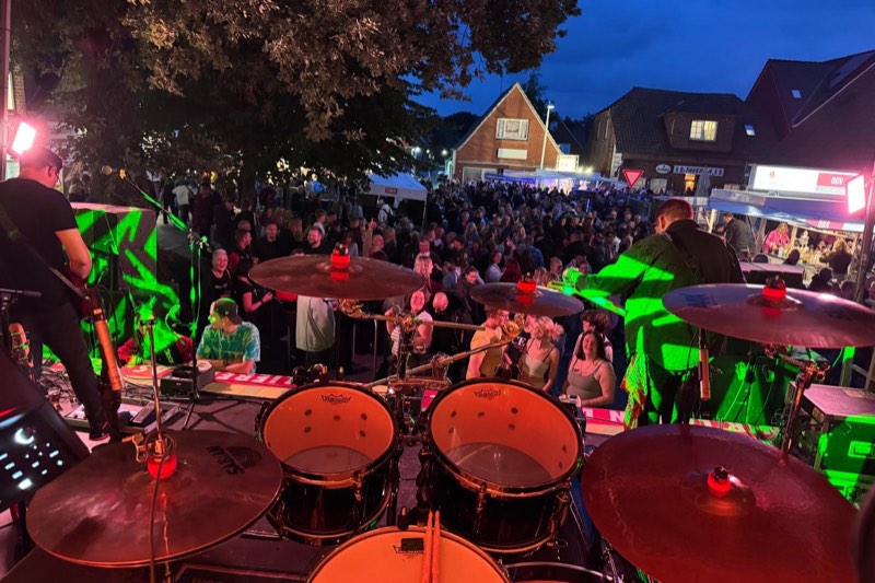 Blick von der Bühne über das Schlagzeug hinweg auf die dicht gefüllte Hauptstraße in der Abenddämmerung, links und rechts Gitarristen von RockShots, im Hintergrund Häuser und Festbuden.