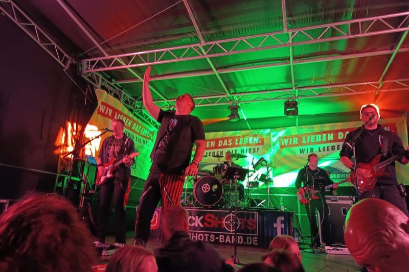 Die Band RockShots spielt im grünen und roten Scheinwerferlicht, der Sänger reckt einen Arm in die Höhe, dahinter Banner von Radio Nordseewelle.