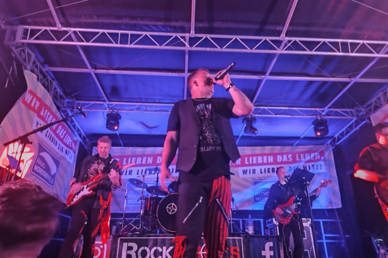 Der Sänger von RockShots singt im blauen Bühnenlicht, hinter ihm Gitarrist, Schlagzeug und Bassist vor Bannern von Radio Nordseewelle.